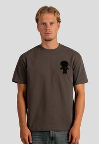 Camiseta de algodón gris con un gráfico texturizado de una muñeca negra en el lado izquierdo del pecho, con un pequeño corazón rojo en el pecho de la muñeca.