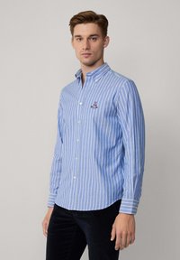 Camisa de botones en azul claro con rayas verticales blancas, que presenta un pequeño logo en el pecho, mangas largas y un dobladillo curvado.