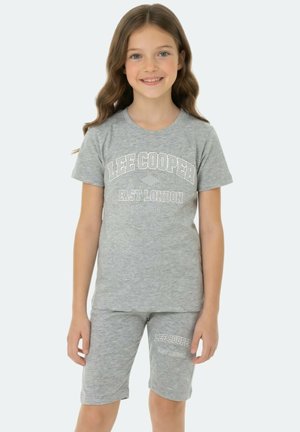 Jeune fille souriante, portant un t-shirt et un short assortis gris clair Lee Cooper avec des lettres blanches, debout devant un arrière-plan uni.