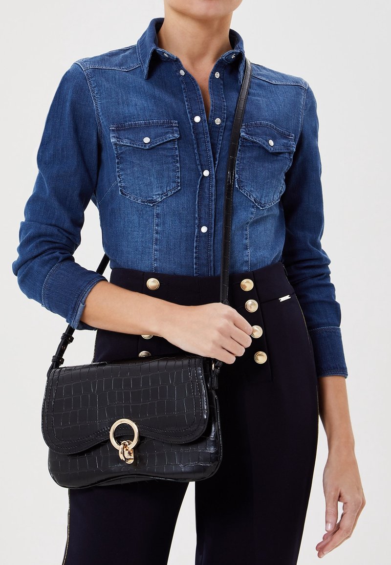 Borsa a tracolla in pelle nera effetto coccodrillo con dettaglio in metallo dorato a forma di anello, forma rettangolare e tracolla. Indossata con una camicia di jeans.
