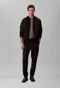 Noor mees seisab ühevärvilise tausta ees, seljas must jakk, hall särk, mustad püksid ja mustad vabaajajalatsid, käed lõdvalt külgedel.