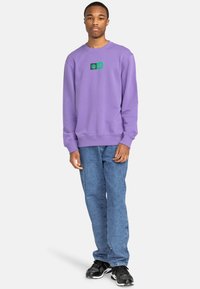Purple sweatshirt with a crew neckline, featuring green graphic patches on the chest. Worn with blue denim jeans and black sneakers.