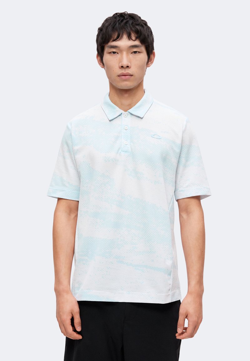 Homme portant un polo bleu clair et blanc à manches courtes avec un motif subtil et un pantalon noir, debout devant un fond uni.