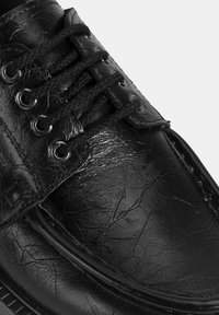 Schwarzer Lederschuh mit strukturierter Oberfläche, runder Zehenform, schwarzen Schnürsenkeln durch silberne Ösen und genähten Details entlang der Sohle.