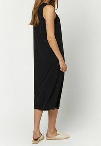 Femme debout dans une robe midi noire sans manches et des espadrilles comfortables beiges, sur fond blanc uni.