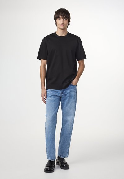 T-shirt en coton noir à manches courtes, associé à un jean en denim bleu clair. Le mannequin se tient les mains dans les poches, portant des chaussures noires.