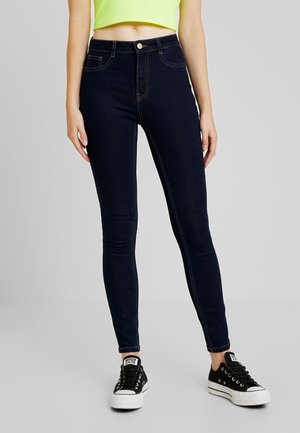 Person iført højttaljede mørkeblå skinny jeans, cropped neongul top og sorte lave sneakers, stående mod en ensfarvet baggrund.