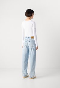 Jeans larges bleu clair à taille haute, avec deux poches arrière et une étiquette en cuir beige sur la ceinture. Haut blanc à manches longues.