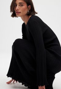 Melna ribētā kleita ar V veida kakla izgriezumu, izplestām piedurknēm un teksturētu audumu. Iezīmē viļņotu apakšmalas apdari un pieguļošu siluetu.