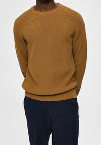 Hnědý žebrovaný pletený svetr s kulatým výstřihem a raglánovými rukávy, v kombinaci s tmavě modrými kalhotami s texturovaným materiálem.
