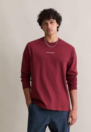 Bordeaux langlevenshirt met ribgebreide manchetten, voorzien van een witte logo-tekst op de borst. Gecombineerd met blauwe denim broek.