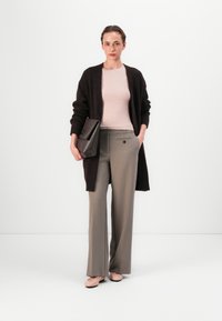 Dunkelbraune offene Strickjacke, hellrosa enges Top, graue Hose mit hoher Taille und Taschen sowie blassrosa Ballerinas. Hält eine strukturierte braune Tasche.