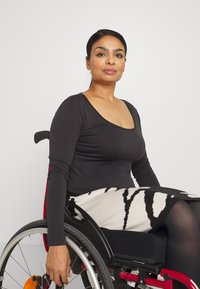 Zwarte blouse met lange mouwen, zwarte en witte gestreepte rok, zittend in een rolstoel met zilveren wielen en een rode frame. Eenvoudige achtergrond.