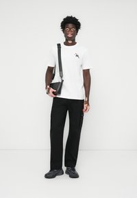 Weißes T-Shirt mit einem kleinen Grafikdesign, schwarze Cargo-Hosen und schwarze Umhängetasche. Die Sneakers sind strukturiert und dunkel. Einfaches Design.