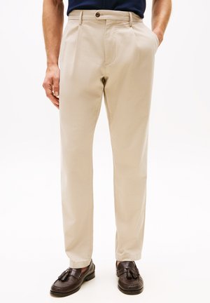 Mann in beigen Hosen und dunklen braunen Quastenmokassins, mit einer Hand in der Tasche, stehend vor einem schlichten weißen Hintergrund.