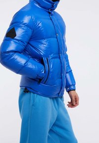 Niebieska kurtka puchowa o połyskującej powierzchni, z kieszeniami na zamek, wysokim kołnierzem oraz czarną naszywką na ramieniu. Połączona z jasnoniebieskimi spodniami.