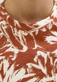 Camiseta de manga corta con cuello redondo, hecha de algodón en color óxido con un patrón de hojas en crema, creando un contraste visual texturizado.