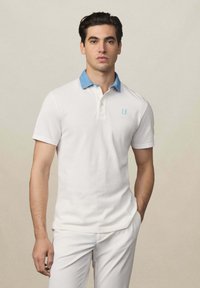 Weißes Poloshirt mit einem denimblauen Kragen und einem kleinen hellblauen Logo. Kurze Ärmel und taillierte Passform. Glattes Baumwollmaterial.
