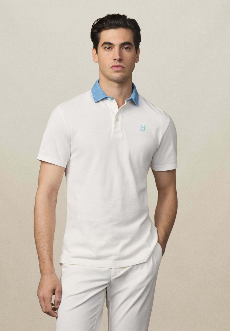 Weißes Poloshirt mit einem denimblauen Kragen und einem kleinen hellblauen Logo. Kurze Ärmel und taillierte Passform. Glattes Baumwollmaterial.