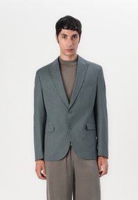 Grå skräddarsydd blazer i ett texturerat tyg, med kavajslag, två knappar och klafffickor, stylad över en brun polotröja.
