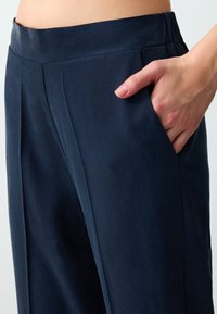 Close-up van de taille en hand van een persoon die in de zakken van een marineblauwe broek met geplooid voorpand rust.
