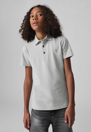 Ljusgrå polo med krage, tre knappar och en liten logotyp. Bärs med mörkgrå denimjeans. Lockigt hår synligt.