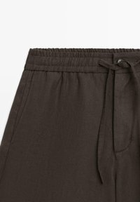 Pantaloni di lino marrone scuro con cintura elastica, tasche laterali, bottone frontale e cintura in tessuto annodata.