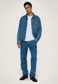 Chaqueta de denim en azul con cierre de botones, bolsillo en el pecho con un logo, combinada con jeans de denim azul a juego y botas negras.
