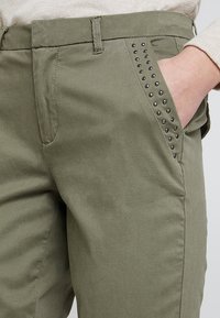 Zielone oliwkowe spodnie o gładkiej fakturze. Posiadają dekoracyjne metalowe ćwieki wzdłuż krawędzi przedniej kieszeni oraz klasyczny prosty krój.