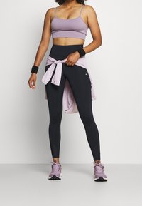 Czarne legginsy o teksturowanym wykończeniu, zestawione z jasnofioletowym stanikiem sportowym i zawiązanym swetrem, uzupełnione fioletowymi butami sportowymi i opaskami na nadgarstki.