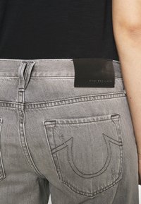 Vaqueros de mezclilla gris con una etiqueta de cuero negra, que presentan bolsillos traseros distintivos con costuras bordadas y una textura suave.
