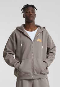 Sudadera gris con cremallera hecha de material suave, con un bolsillo canguro, capucha con cordón y un logo naranja en el lado izquierdo del pecho.