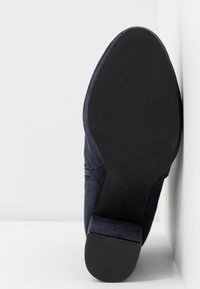 Semelle noire d'une chaussure à talon bleu foncé reposant verticalement contre un angle blanc entre un mur et le sol.