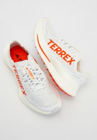 White athletic shoes with a mesh upper, featuring orange "TERREX" branding, red insoles, and a textured white midsole and orange outsole.