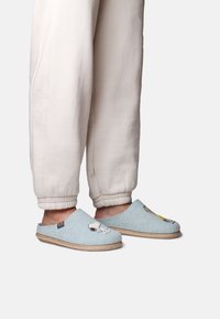 Pantofole di feltro azzurro chiaro con design di cartoni animati, caratterizzate da una texture morbida e suola in fibra naturale, abbinate a pantaloni jogger di colore crema.
