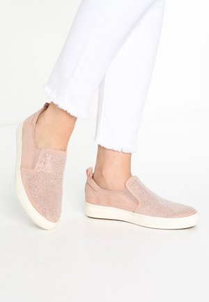 Person iført hvide bukser med frynset kant og lyserøde slip-on sneakers med rhinestenspynt på hvid baggrund.