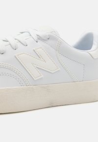 White leather sneakers with a textured cream rubber sole, featuring a prominent raised "N" logo on the side and thin white laces.
