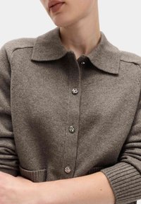 Brązowy kardigan wełniany z kołnierzykiem, z zapięciem na srebrne guziki i ściągaczami przy mankietach. Posiada boczne kieszenie i miękką teksturę.