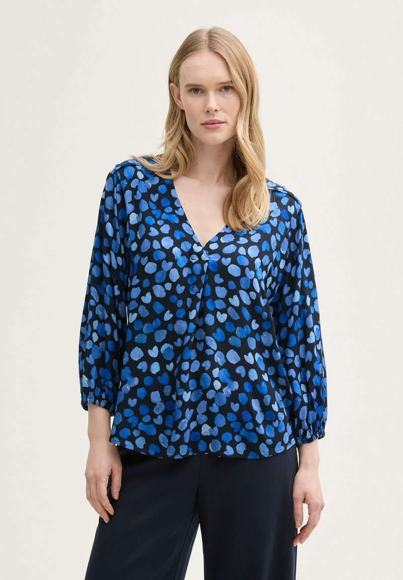 Donna che indossa una blusa a maniche lunghe blu e nera a pois con scollo a V e pantaloni neri larghi, in piedi contro uno sfondo semplice.