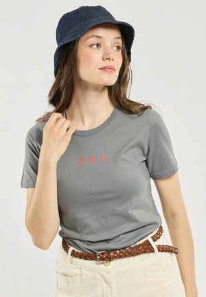 Femme portant un t-shirt gris avec un motif rouge, un pantalon crème, une ceinture tressée marron et un chapeau seau bleu foncé, regardant sur le côté.