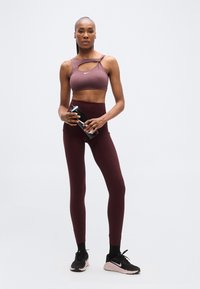 Top sportivo color malva con design con ritagli, abbinato a leggings a vita alta color amaranto. Scarpe sportive nere con suola rosa. Tiene una bottiglia d'acqua nera.