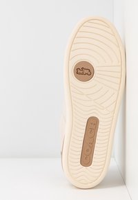 Semelle de basket beige montrant un motif d'adhérence, un emblème rond avec une calèche tirée par des chevaux, et le nom de la marque "COACH" en lettres en relief.