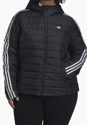 Personne portant une veste matelassée noire avec des bandes blanches sur les manches et un logo Adidas blanc sur la poitrine, zippée.