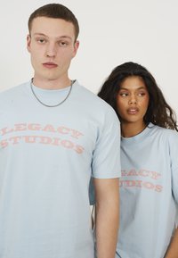 Light blue cotton t-shirts with crew necks, featuring pink vintage-style "LEGACY STUDIOS" text across the front. Simple, relaxed fit design.