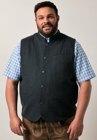 Mørk uldvest med høj krave, der har knaplukning, to lommer og en struktureret overflade, båret over en blå gingham-skråternet skjorte.