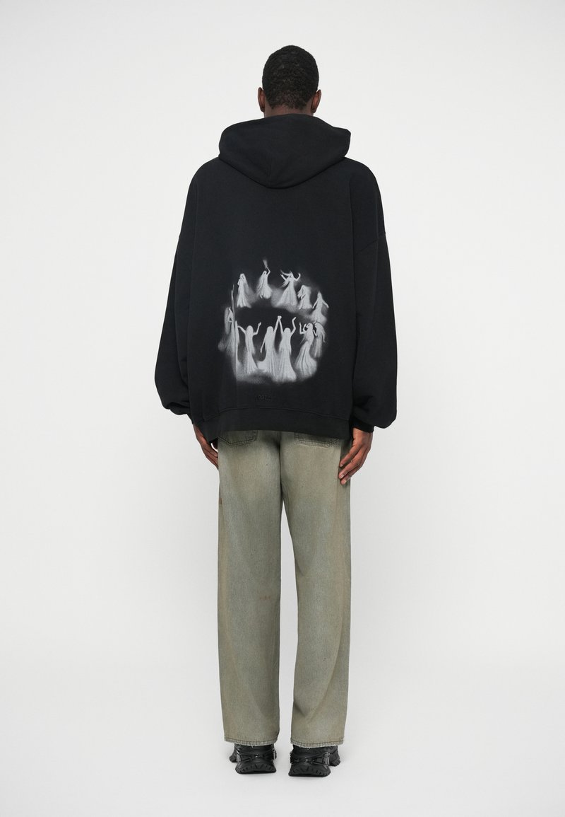 Man in een zwarte hoodie met witte spookfiguren die hand in hand in een cirkel staan op de achterkant, gecombineerd met lichtgrijze jeans en zwarte schoenen.