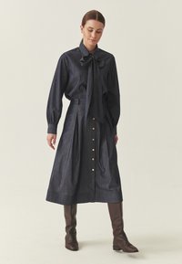 Kobieta ubrana w ciemnoniebieską midi sukienkę z denimu z kołnierzem na kokardę i złotymi guzikami, zestawioną z brązowymi skórzanymi kozakami za kolano.