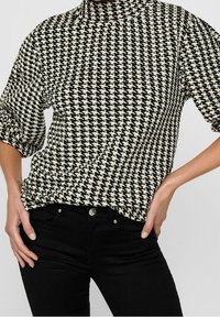 Haut à motif pied-de-poule noir et crème avec un col haut et des manches courtes bouffantes. Le tissu semble texturé. Porté avec un pantalon noir.