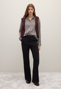 Chemise boutonnée rayée noire et blanche associée à un pantalon noir taille haute et un gilet en tricot bordeaux foncé, porté sur les épaules.
