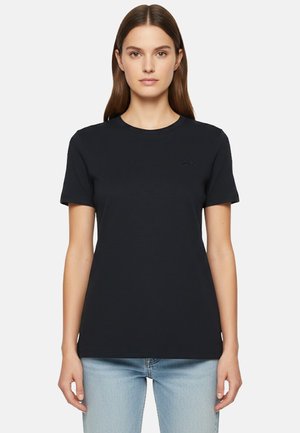 Donna con capelli castani lisci che indossa una t-shirt nera a maniche corte e jeans azzurri, in piedi contro uno sfondo bianco.